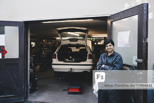 Portrait einer selbstbewussten Mechanikerin mit gekreuzten Armen am Eingang der Autowerkstatt