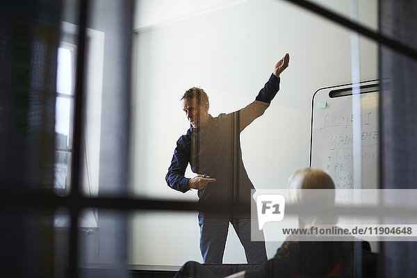 Businesswoman looking at male professional giving presentation against wall in board room seen from glass at creative of