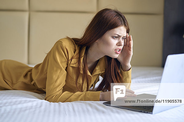 Frustrierte kaukasische Frau liegt auf dem Bett und benutzt einen Laptop