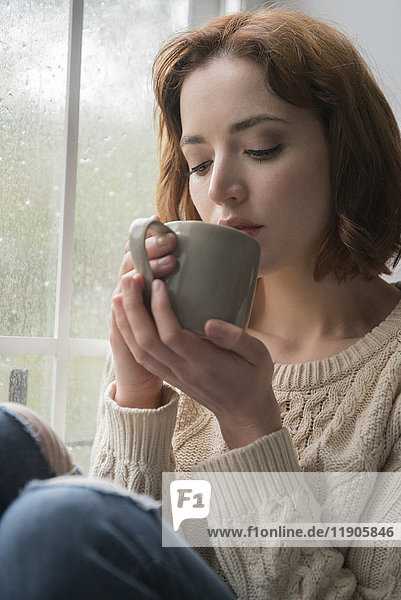 Kaukasische Frau trinkt Kaffee in der Nähe eines regnerischen Fensters