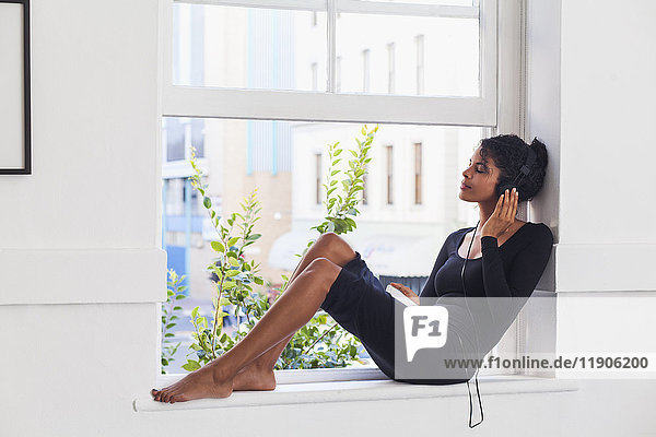 Mixed-Race-Frau sitzt auf der Fensterbank und hört Kopfhörer