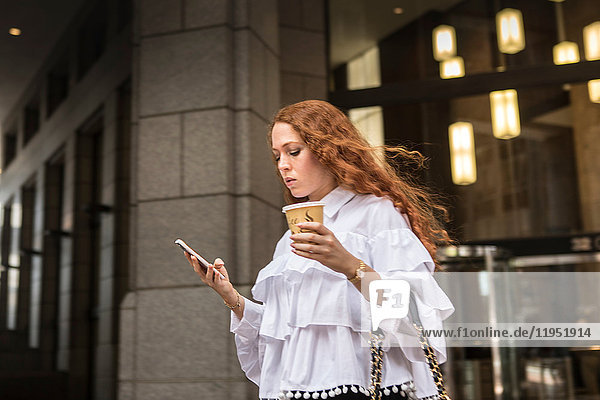 Junge Geschäftsfrau mit Kaffee zum Mitnehmen mit Blick auf Smartphone auf dem Bürgersteig  New York  USA