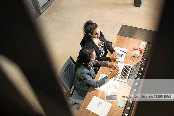 Two businesswomen sitting at desk  using laptop  looking at smartphone  elevated view