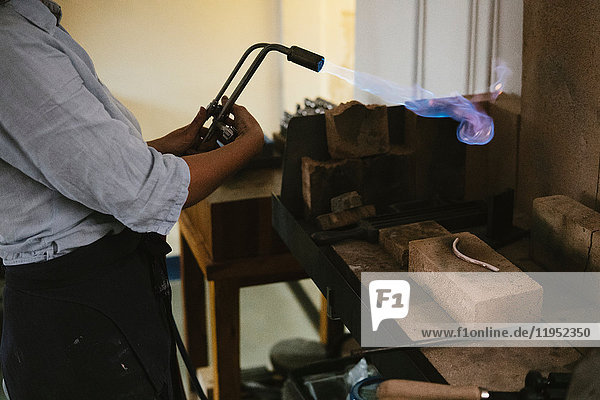 Mid section of female jeweller using flaming blow torch at workbench