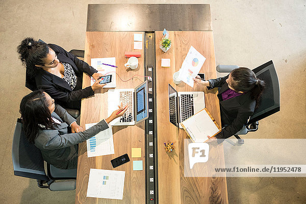 Drei Geschäftsfrauen sitzen am Schreibtisch  benutzen Laptops  halten ein Smartphone in der Hand  Blick nach oben