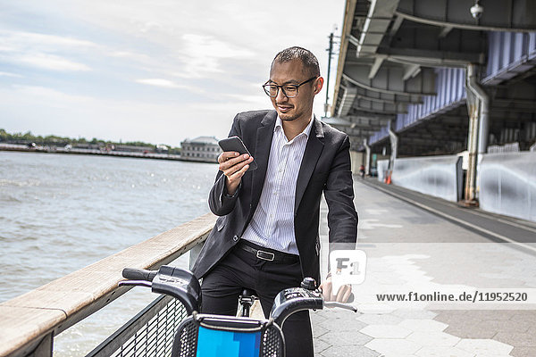 Geschäftsreisender Radfahrer betrachtet Smartphone am Wasser  New York  USA