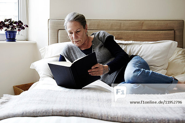 Senior woman with grey hair reclining on bed reading book