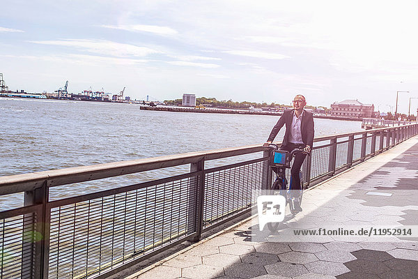 Mittelgroßer erwachsener Geschäftsmann mit dem Fahrrad entlang der Uferpromenade des Stadtflusses