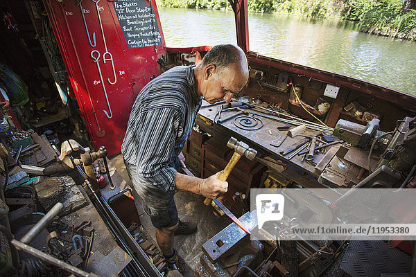 Hochwinkelaufnahme eines Schmiedes an seiner Werkbank auf seinem Arbeitsboot auf dem Wasser  der auf heißes Metall hämmert.
