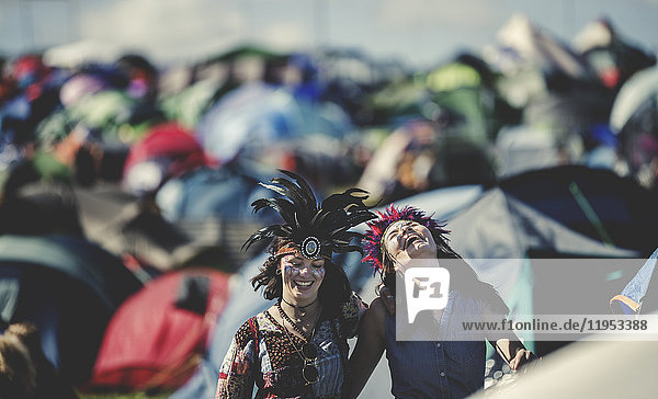 Zwei lächelnde junge Frauen bei einem sommerlichen Musikfestival  mit Federkopfschmuck bemalt  stehen zwischen den aufgeschlagenen Zelten.