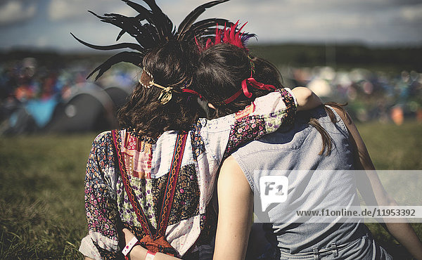 Rückansicht von zwei jungen Frauen bei einem Sommer-Musikfestival mit Federkopfschmuck  den Arm um die Schulter gelegt.