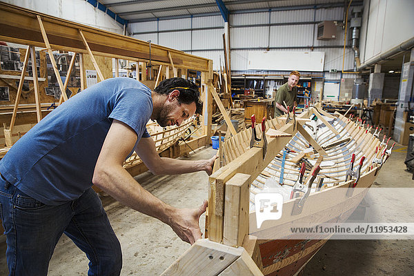 Zwei Männer in der Werkstatt eines Bootsbauers  die gemeinsam an einem hölzernen Bootsrumpf arbeiten.