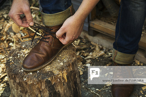 Ein Mann in Denim-Jeans mit Umschlägen sitzt angelehnt  um die Schnürsenkel seiner braunen Lederschuhe zu binden  einen Fuß hoch auf einem Spaltblock.