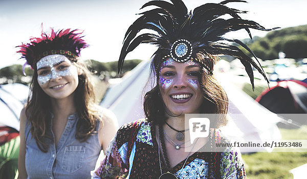 Zwei lächelnde junge Frauen bei einem Sommer-Musikfestival mit bemaltem Gesicht und Federkopfschmuck zwischen Zelten stehend.