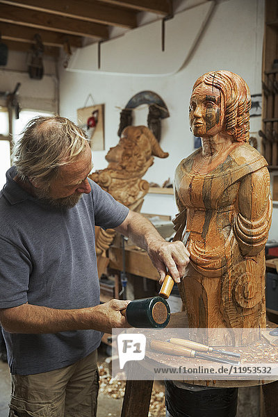 Ein Holzschnitzer  der in seiner Werkstatt steht und mit Handwerkzeugen  Hammer und Meißel eine hölzerne weibliche Schiffsgestalt aus Holz formt und verziert.
