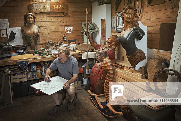 A craftsman  woodworker seated on a stool in a workshop working on a drawing  sketching using charcoal. Surrounded by wooden carved and painted female ship's figureheads.