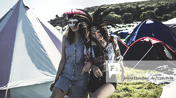 Zwei junge Frauen bei einem Sommer-Musikfestival bemalte Gesichter  Federkopfschmuck tragend  Arm in Arm zwischen den Zelten spazierengehend.