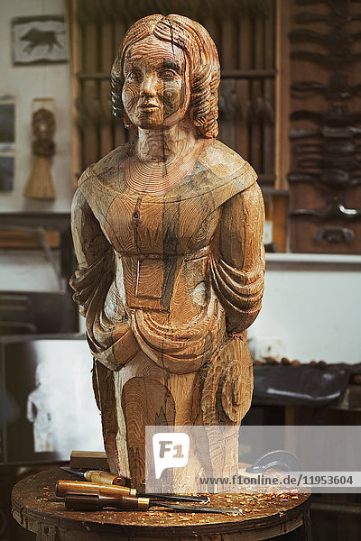 Eine geschnitzte hölzerne weibliche Schiffsgestalt auf einer Bank in einer Werkstatt mit einem Muster aus Holzmaserungsmarkierungen