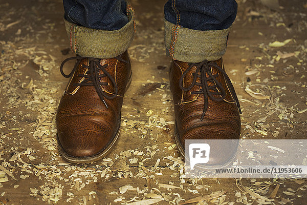 Füße und Knöchel eines Mannes in Denim-Jeans mit Umschlag und traditionellen braunen geschnürten Lederschuhen stehen zwischen Holzspänen auf einem Werkstattboden.