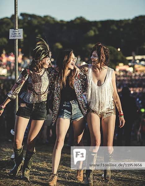 Drei junge Frauen bei einem Sommer-Musikfestival in Hotpants und Wellington-Stiefeln  mit Federkopfschmuck und gemalten Gesichtern  die Arme um die Schulter gelegt und lächelnd.