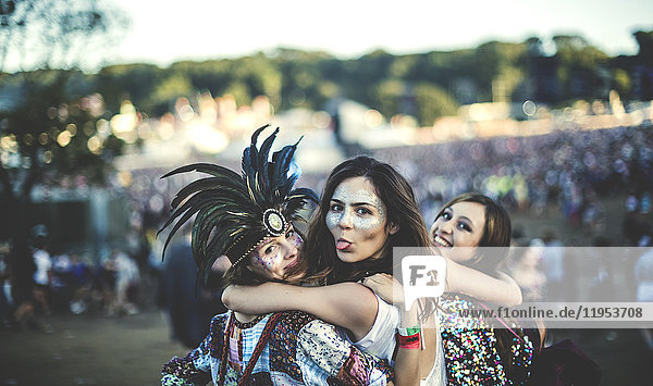 Drei junge Frauen bei einem Sommer-Musikfestival Federkopfschmuck und bemalte Gesichter  lächeln in die Kamera und strecken ihre Zunge heraus.
