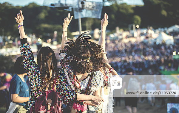Rückansicht von drei jungen Frauen  die nebeneinander bei einem Sommer-Musikfestival mit Federkopfschmuck und erhobenen Armen stehen.