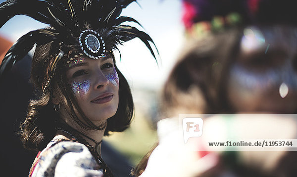 Junge Frau bei einem Sommer-Musikfestival mit geschminktem Gesicht  Federkopfschmuck und Blick in die Kamera.