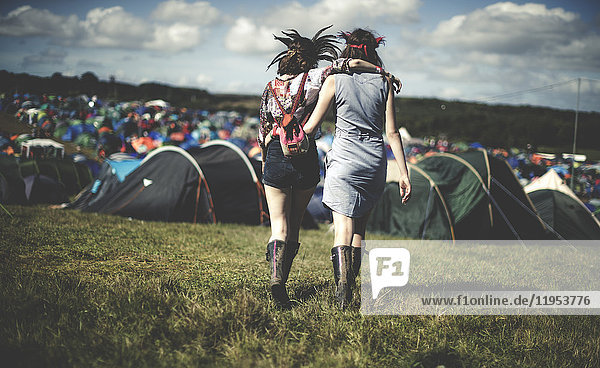 Rückansicht von zwei jungen Frauen bei einem Sommer-Musikfestival mit Federkopfschmuck  die Arm in Arm auf Zelte zugehen.