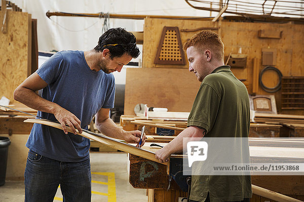 Two men standing at a workbench in a boat-builder's workshop  working on piece of wood.