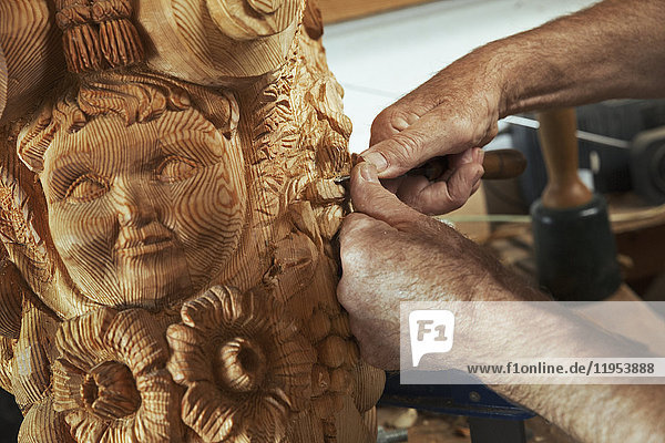 Nahaufnahme eines Kunsthandwerkers  eines Holzschnitzers  der in einer Werkstatt an einer hölzernen Galionsfigur mit menschlichem Gesicht und floralen Formen arbeitet und einen kleinen Meißel hält.