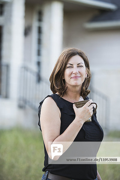 Eine Frau in einem ärmellosen Hemd  die einen Becher in der Hand hält  steht vor einem Landhaus.