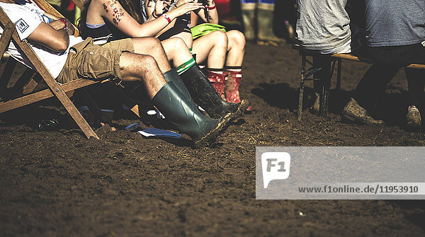 Menschen  die auf Liegestühlen im Schlamm sitzen und Wellington-Stiefel tragen  bei einem Sommer-Musikfestival.