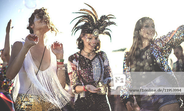 Drei lächelnde junge Frauen bei einem Sommer-Musikfestival mit geschminktem Gesicht und Federkopfschmuck  die in der Menge tanzen.