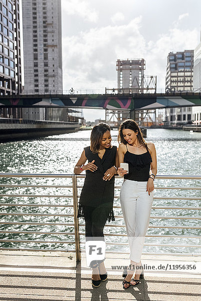 Two women on a bridge sharing a cell phone in the city