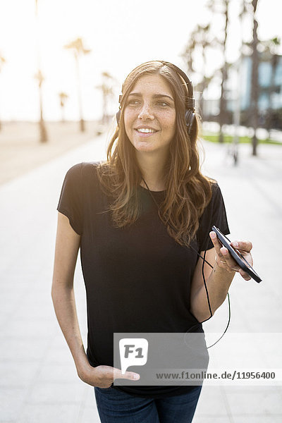 Lächelnde junge Frau an der Strandpromenade beim Musikhören