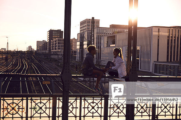 Deutschland  München  Junges Paar auf der Brücke sitzend  Sonnenuntergang genießend