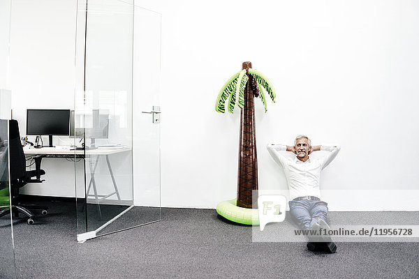 Smiling mature businessman sitting next to inflatable palm tree in office