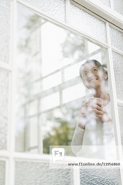 Lächelnde Frau mit einer Tasse Kaffee,  die aus dem Fenster schaut.