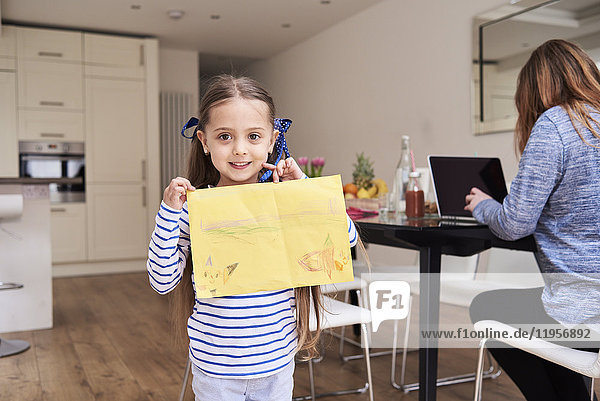 Porträt eines lächelnden Mädchens mit Zeichnung  während ihre Mutter im Hintergrund am Laptop arbeitet.