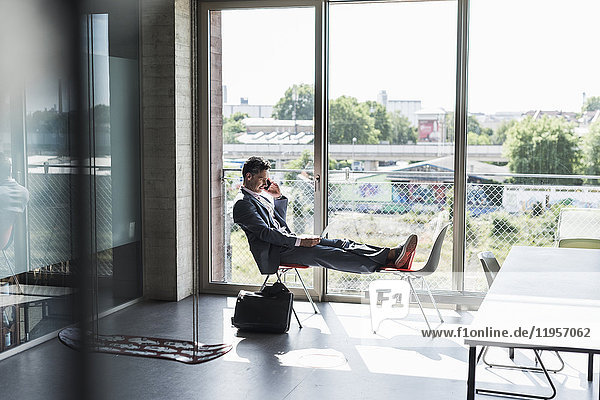 Geschäftsmann am Fenster sitzend mit erhobenen Füßen  am Laptop arbeitend und telefonierend