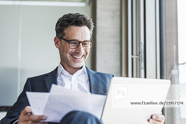 Portrait of laughing businessman with documents looking at tablet