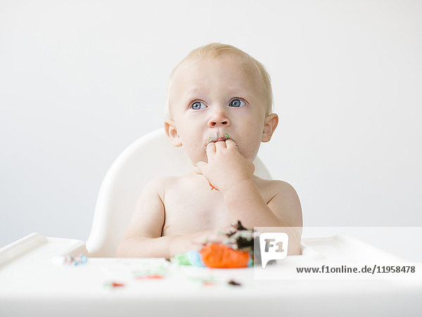 Baby boy (12-17 months) sitting in high chair and eating cupcake
