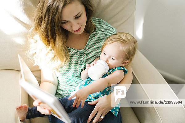 Mutter mit kleinem Mädchen (12-17 Monate) sitzt im Wohnzimmer und liest ein Buch