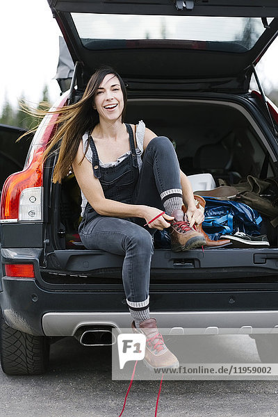 Lächelnde Frau  die im Kofferraum eines Autos sitzt und Wanderschuhe bindet