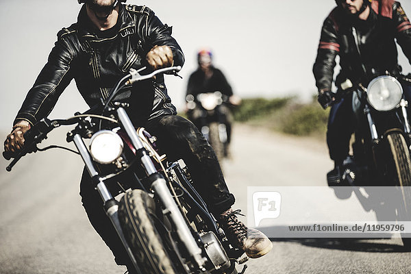 Three men wearing leather jackets riding cafe racer motorcycles along rural road.