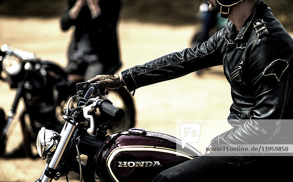 Seitenansicht eines Mannes in schwarzer Lederjacke  der auf einem Cafe-Rennmotorrad auf einer staubigen Schotterstraße sitzt.