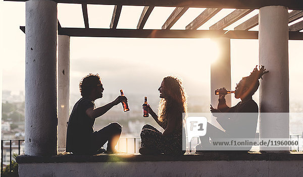 Silhouette von zwei Männern und einer Frau  die bei Sonnenuntergang im Freien unter einer Pergola sitzen und Bierflaschen halten.