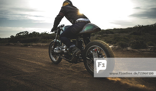 Rear view of man wearing crash helmet riding cafe racer motorcycle on a dusty dirt road.