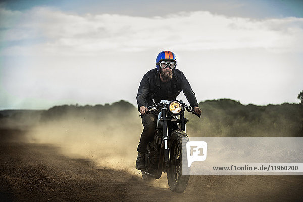Man wearing open face crash helmet and goggles riding cafe racer motorcycle on a dusty dirt road.