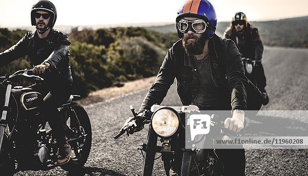 Three men wearing open face crash helmets and goggles riding cafe racer motorcycles along rural road.